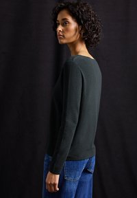Maglione a maglia verde scuro con collo rotondo e maniche lunghe, abbinato a jeans blu. La texture è a coste. Lo sfondo è nero.
