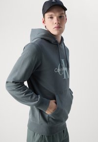 Siva bombažna kapuca z sprednjim žepom in kapuco na vrvico, s svetlo obarvanim logotipom "Calvin Klein" natisnjenim na prsih.