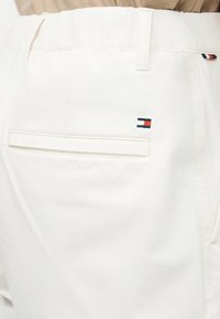 Κοντινό πλάνο της πίσω τσέπης λευκών παντελονιών με το λογότυπο Tommy Hilfiger σε μπλε ναυτικό, κόκκινο και λευκό, και θηλιά ζώνης, φορεμένα με μπεζ τοπ.