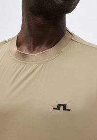 Detailný záber osoby nosiacej béžové tričko s okrúhlym výstrihom a malým čiernym geometrickým logom na hrudi.