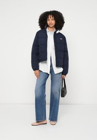 Tamsiai mėlynas pūkinis švarkas su užtrauktuku, dėvimas ant balto marškinių su sagomis. Šviesiai mėlynos plačios kelnės ir lygūs batai. Juoda rankinė.