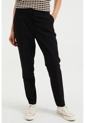 Person iført sorte, tætsiddende bukser og hvide sneakers, stående med den ene hånd i lommen mod en ensfarvet baggrund.