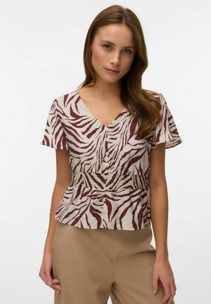 Vrouw die een kortermige V-hals blouse draagt met een bordeauxrode en witte zebraprint, knopen aan de voorkant, en beige hoge taille broek, staand tegen een witte achtergrond.