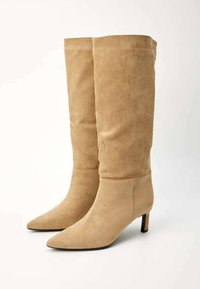 Bottes en suède beige montantes avec un bout pointu et un talon fin, arborant une texture lisse et une silhouette épurée.