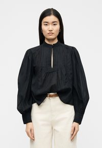 Γυναίκα με μακριά ίσια μαύρα μαλλιά, που φοράει μαύρο μπλουζάκι με φουσκωτά μανίκια και κρεμ ψηλόμεσο παντελόνι με καφέ ζώνη.