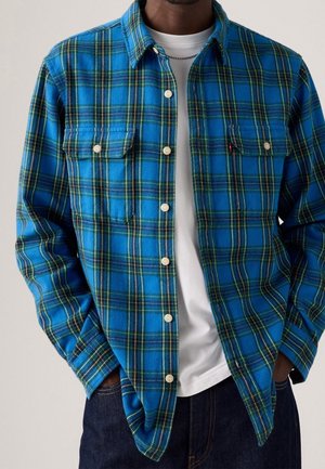 Homme portant une chemise à carreaux bleu et vert boutonnée sur un t-shirt blanc avec un jean foncé et les mains dans les poches.