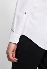 Camicia bianca con una texture liscia, polsini con bottoni e un dettaglio ricamato di una rosa nera sul polsino sinistro. Pantaloni neri parzialmente visibili.