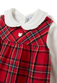 Vestido de cuadros rojos con cuello blanco, presenta una falda plisada y un pequeño detalle de corazón dorado en el corpiño. Textura de tela suave.