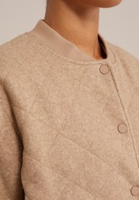 Beige gewatteerde jas van zacht, textuur materiaal, met een geribbelde kraag en twee ronde knopen voor sluiting.