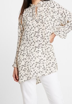 Woman wearing a cream long-sleeve blouse with black floral print, ruffled collar, tie detail, and white pants.