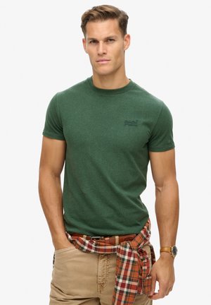 Green cotton t-shirt with short sleeves and a crew neckline. Features a subtle logo on the chest, paired with a plaid shirt tied at the waist.