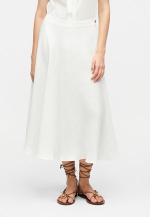 Naine kannab valget voolavat midi-seelikut, mida täiendab pruunide nahast nöörtega sandaalide koos kuldsete rõngaste aksentidega.