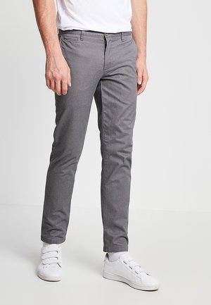 Mężczyzna w obcisłych szarych spodniach, białych skarpetkach, białych trampkach na rzepy i białej koszuli, stoi na tle jasnego, jednolitego tła.