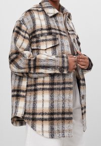 Flanelová košile s kostkovaným vzorem v béžové, černé a bílé barvě. Dlouhé rukávy, zapínání na knoflíky a dvě přední kapsy. Měkká textura.