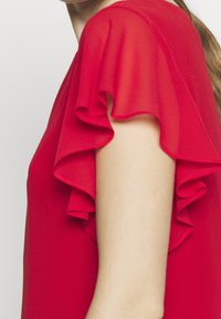 Blouse sans manches rouge avec un détail de tissu superposé et froncé autour du bras et de l'épaule, portée par une personne à la peau claire.