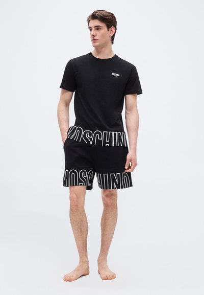 Νεαρός άνδρας που στέκεται ξυπόλυτος φορώντας μαύρο t-shirt και σορτς Moschino με έντονα λευκά γράμματα λογότυπου στο τελείωμα, μπροστά σε απλό φωτεινό φόντο.