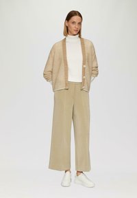 Beige Strickjacke mit subtiler gestreifter Textur, ausgestattet mit drei Knöpfen. Kombiniert mit einem cremefarbenen Rollkragenpullover und weiten beigen Hosen. Weiße Sneaker.