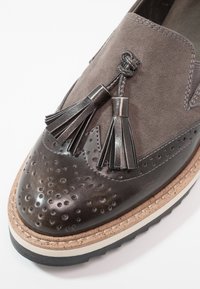 Mocassin marron en cuir et daim avec des détails perforés, deux pampilles en cuir, semelle superposée contrastante et surpiqûres visibles sur le bord supérieur.
