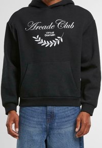 Černá mikina s kapucí vyrobená z měkkého materiálu, s bílým textem "Arcade Club" a "Virtual Domain" a designem listu nad přední kapsou.