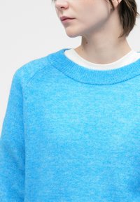 Asmuo, vilkintis ryškiai mėlyną megztinį per baltą marškinį, žiūri į šoną prieš paprastą baltą foną.