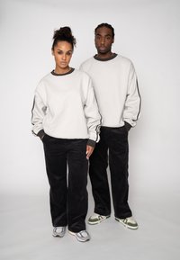 Hanorace gri din bumbac cu manșete negre ribbate, asortate cu pantaloni negri din catifea cu dungi. Ambele poartă adidași în tonuri neutre. Croială simplă și lejeră.