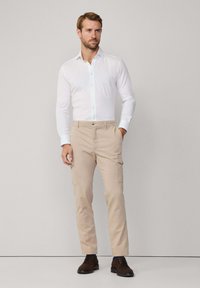 Biała koszula na guziki w zestawieniu z beżowymi spodniami cargo. Spodnie mają boczne kieszenie i zamek błyskawiczny. Ciemnobrązowe buty dopełniają outfit.