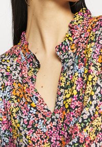 Zwarte bloemenblouse met levendige, veelkleurige patronen, een gerimpelde kraag en een V-halsontwerp. De stof heeft een lichte textuur.