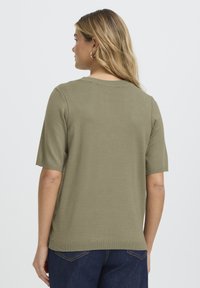 Īsroku olīvu zaļas krāsas adīts tops ar apaļu kakla izgriezumu un ribota auduma faktūru, redzams no aizmugures. Kombinēts ar tumši zilām džinsām.