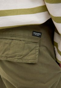 Olivově zelené látkové kalhoty s náprsní kapsou a černou nálepkou s nápisem "JACK & JONES EST. 1990," částečně zakryté pruhovaným topem.