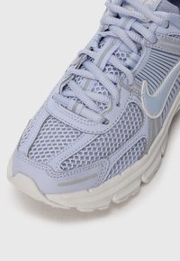 Sapatilha desportiva azul claro com parte superior em malha e acentos sintéticos, apresentando fecho com atacadores, design texturizado e uma sola branca acolchoada.