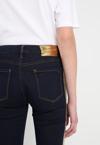 Personne portant un jean bleu foncé avec des bandes latérales dorées et une étiquette marron Tommy Hilfiger sur la ceinture, associée à une chemise blanche.