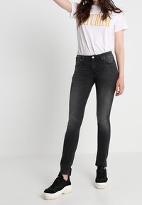 Svarta skinny jeans med en slät textur och midja i normalhöjd, tillsammans med en vit t-shirt med gula bokstäver, och svarta sneakers.