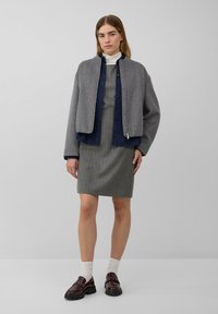 Pelēka vilnas bomber jakas ar rāvējslēdzēju priekšpusē, virs piegulošas pelēkas svītrainas kleitas. Modelis valkā tumši sarkanas loafers un baltas ribotas zeķes.