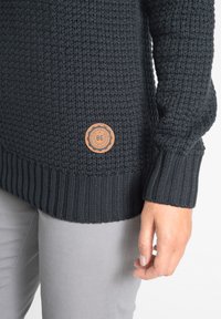 Maglione nero lavorato a maglia con un motivo testurizzato, orlo e polsini a coste. Presenta una patch circolare marrone del marchio in fondo.