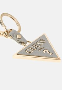 Portachiavi triangolare in materiale grigio chiaro texture con dettagli in metallo dorato; presenta il logo in rilievo "GUESS USA" e un punto interrogativo.