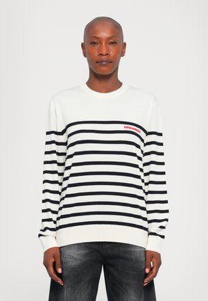 Personne avec la tête rasée portant un pull à rayures horizontales blanches et noires avec le logo rouge "DSQUARED2", associé à un jean foncé.