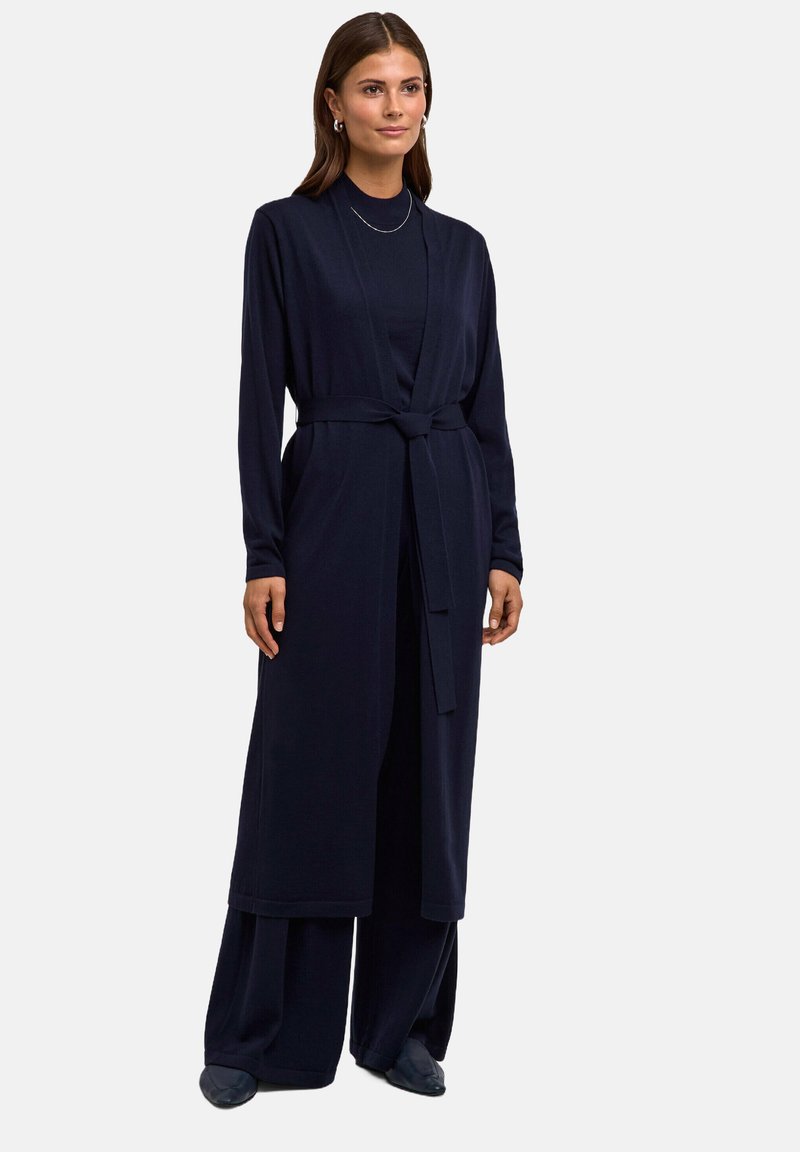 Długi granatowy kardigan, pasek w talii, dopasowane rękawy, wykonany z miękkiego materiału. Noszony na zestaw, który jest w tym samym kolorze, o prostym wzorze.