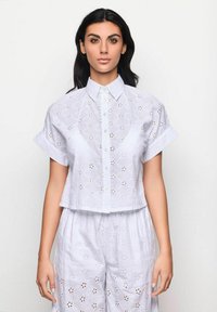 Camicia bianca corta in cotone ricamato, con maniche corte, chiusura frontale a bottoni e disegni floreali ritagliati.