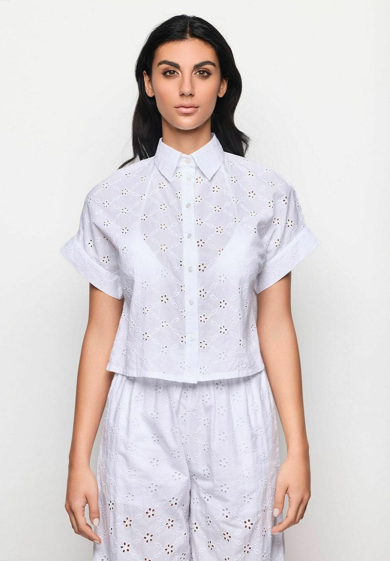 Camicia bianca corta in cotone ricamato, con maniche corte, chiusura frontale a bottoni e disegni floreali ritagliati.