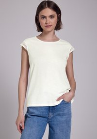 Joven mujer con cabello corto castaño, vistiendo una blusa sin mangas blanca con bordado de encaje y jeans azules, de pie contra un fondo sencillo.