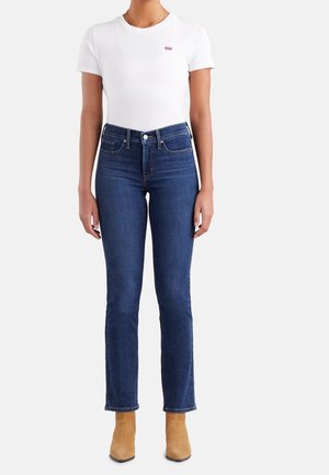 Woman wearing a white short-sleeve t-shirt, dark blue high-waisted jeans, and tan ankle boots standing against a plain background.