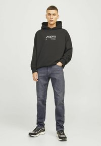 Hoodie noir avec logo sur la poitrine, associé à un jean bleu foncé et des baskets noires avec des accents gris. Style décontracté et design détendu.