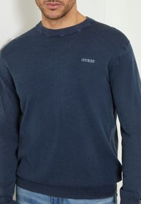 Mann in einem dunkelblauen Langarmpullover mit "GUESS"-Logo auf der Brust und hellblauen Jeans, stehend vor einem weißen Hintergrund.