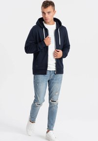 Námořnická mikina na zip s kapucí na šňůrku, kombinovaná s opranými světle modrými džíny a bílými sneakers, nošená přes jednoduché bílé tričko.
