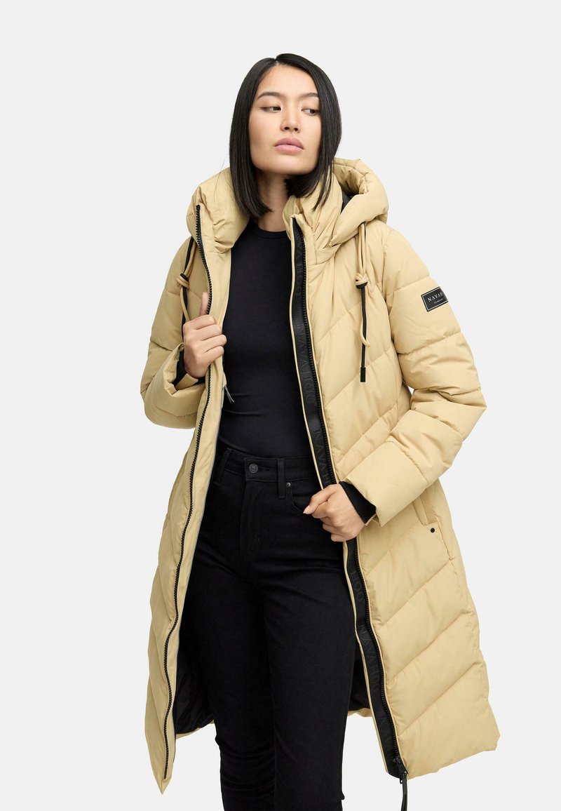 Beige lang dunjakke med hætte, med en kviltet design, frontlynlås og sorte detaljer. Bæres over en sort top og bukser.
