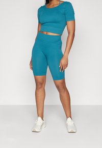 Turkusowy rybnikowy crop top z krótkimi rękawami w zestawie z dopasowanymi, wysokimi szortami rowerowymi. Obuwie składa się z białych butów sportowych.