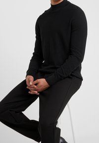 Homme assis sur un tabouret portant un pull noir à col roulé et un pantalon noir, les mains doucement croisées sur les genoux.