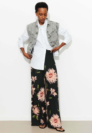 Femme portant une chemise blanche à boutons, un gilet en denim gris sans manches, un pantalon large noir avec un grand imprimé floral rose et des sandales noires.