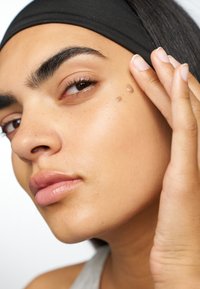 Nærbilde av en jevn, lys brun hudtone med en liten dråpe serum på huden. Subjekter har på seg et svart hodebånd og har buede øyenbryn.