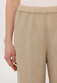 Pantalon en lin beige avec une taille élastique, présentant une texture douce et une coupe décontractée. Une main visible repose à côté du vêtement.
